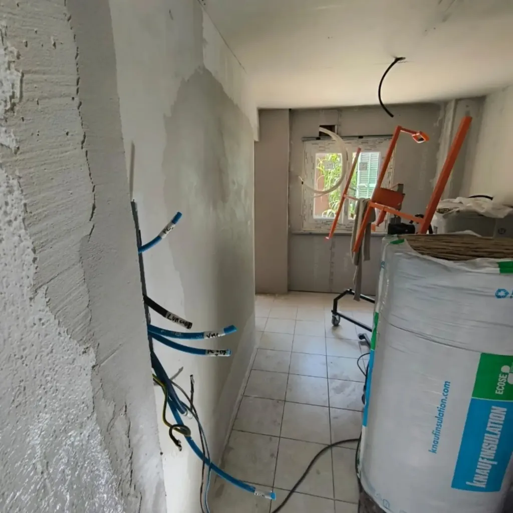 Pièce en rénovation avec tuyaux multicouche bleus encastrés dans un mur enduit, rouleau d'isolant et carrelage ancien.