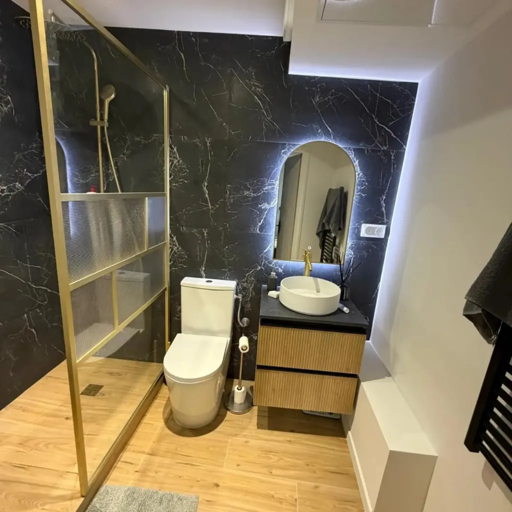 Salle de bain avec mur noir veiné rétroéclairé, paroi de douche à profils laiton, WC et meuble vasque suspendu.