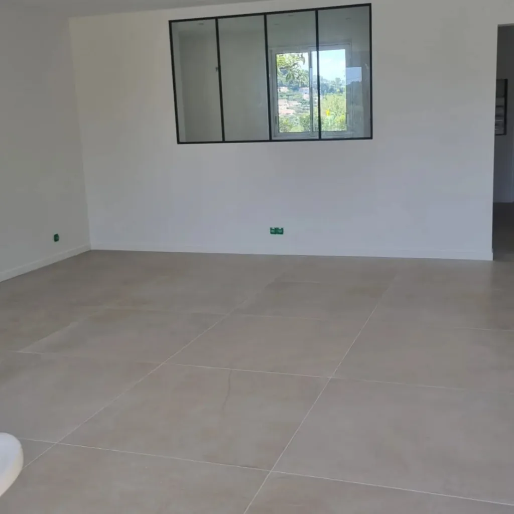 Séjour lumineux vide avec sol en grès cérame gris-beige grand format aux joints fins et plinthes blanches.