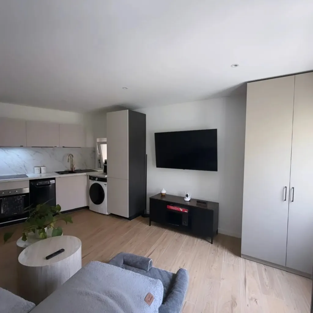 Séjour avec cuisine ouverte beige, parquet clair, télévision murale et grandes armoires, sans occupant.