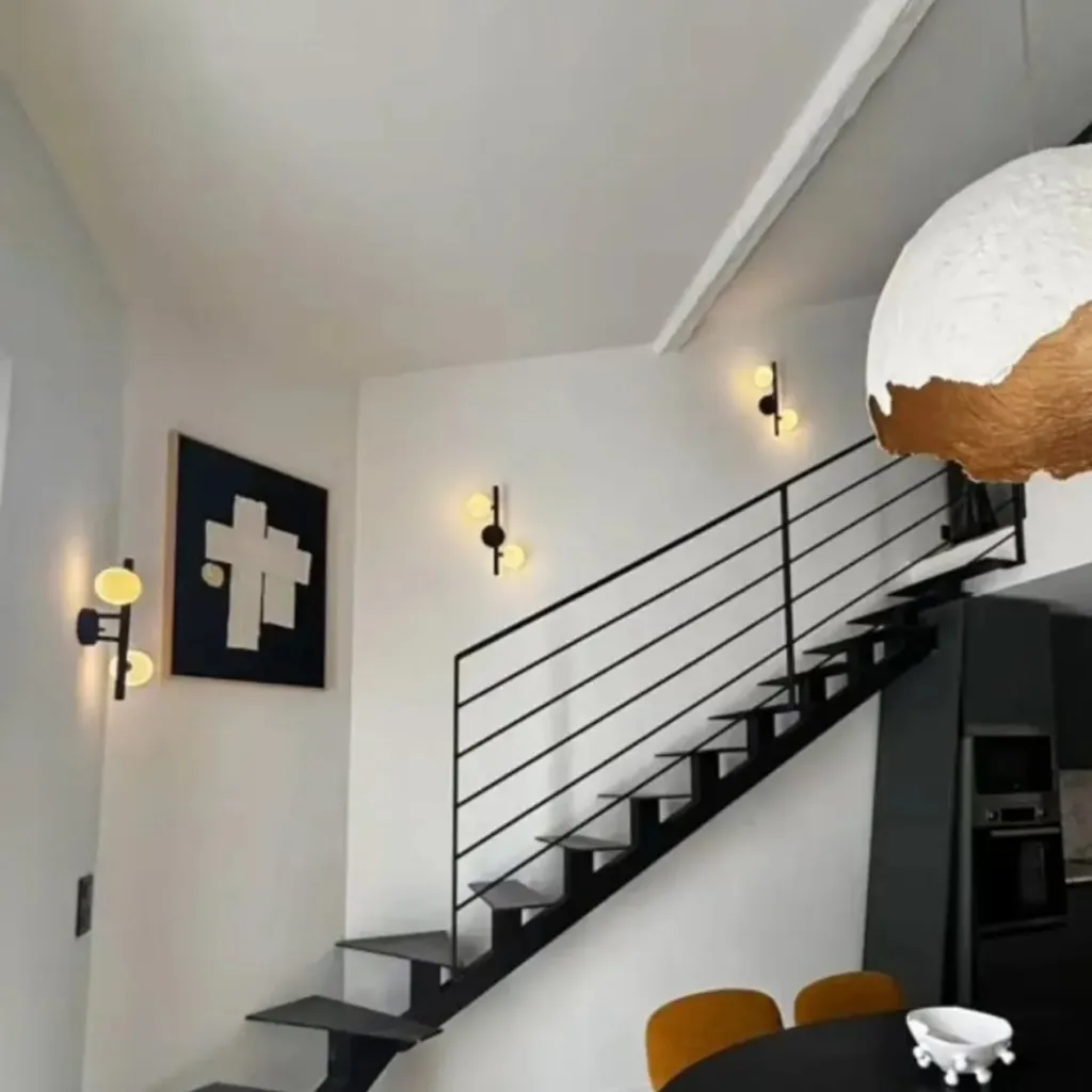 Escalier métallique à marches suspendues, suspension design, table ronde et cuisine noire au fond.