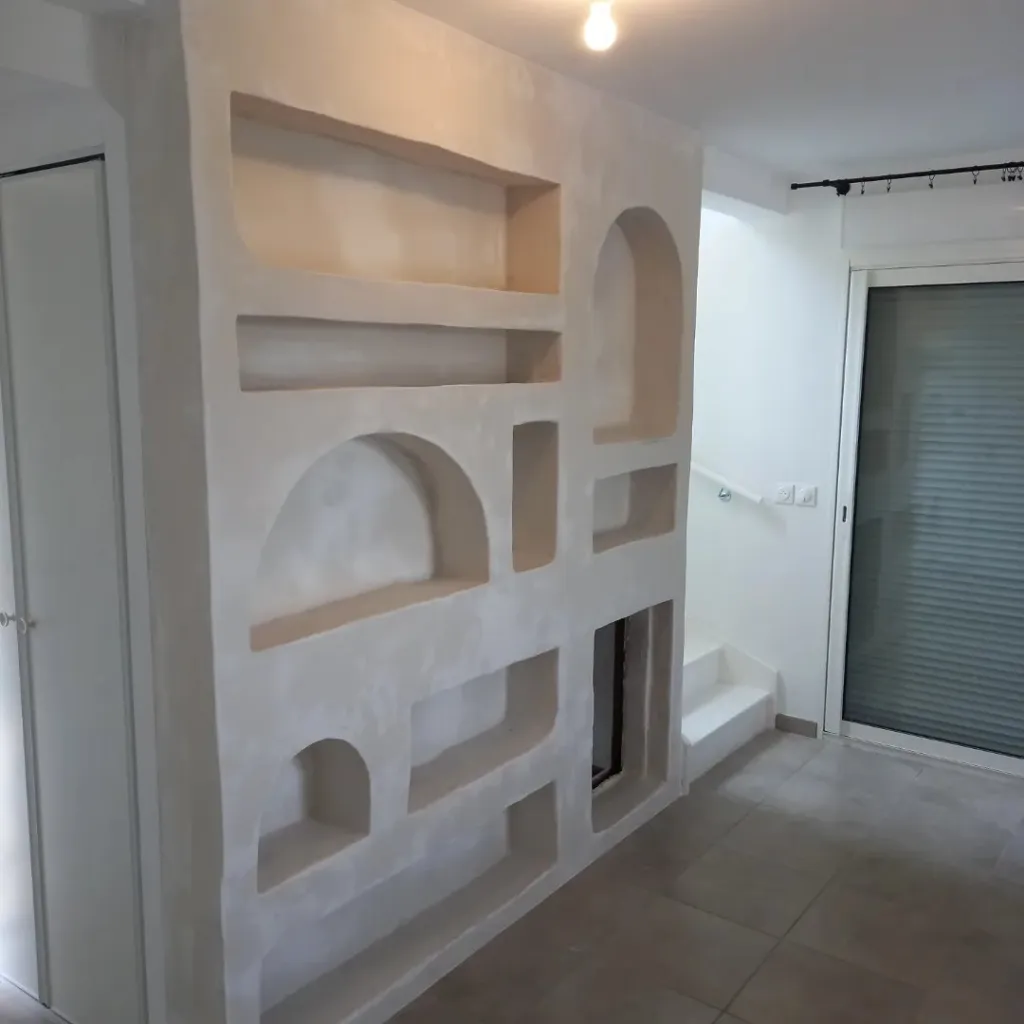 Mur de séparation en plaque de plâtre sculpté avec niches, étagères et arches de tailles variées, dans une entrée près d'un escalier.