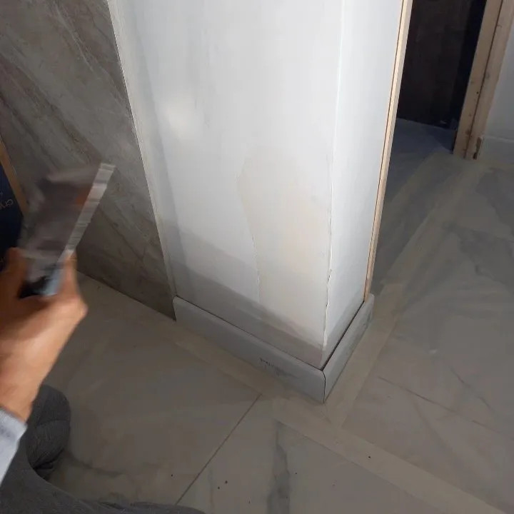 Mains avec couteau à enduire sur un angle de cloison blanche au-dessus d'un sol en marbre protégé par du ruban.