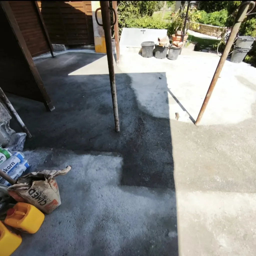Dalle béton en cours de préparation sous un abri bois avec sacs de ciment et jerricane au premier plan.