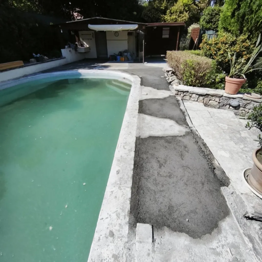 Vue large d'une piscine à margelle blanche avec une bande de chape neuve, abri de jardin au fond.