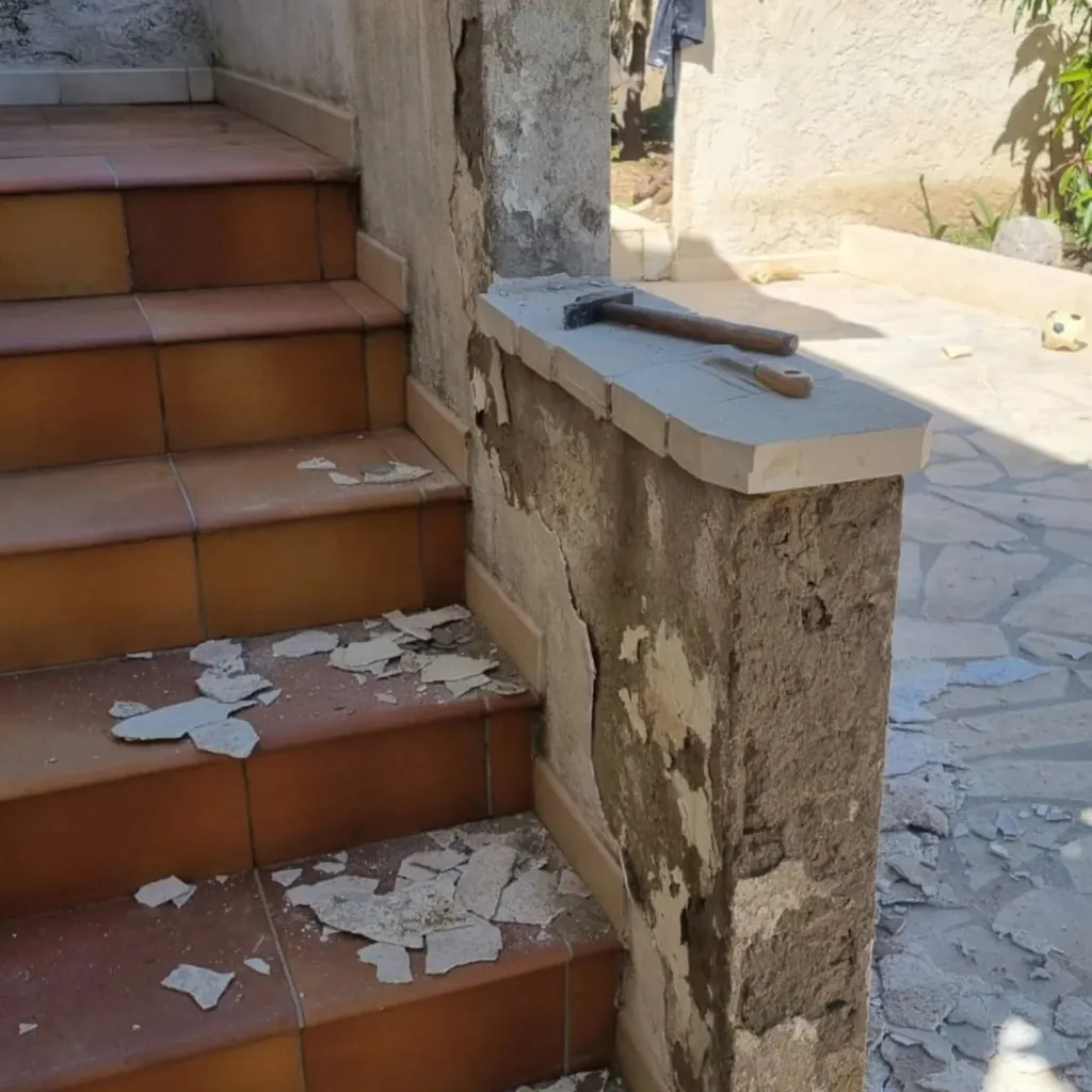 Muret en pierre au crépi dégradé le long d'un escalier carrelé terre cuite, éclats de dalles au sol et marteau posé dessus.