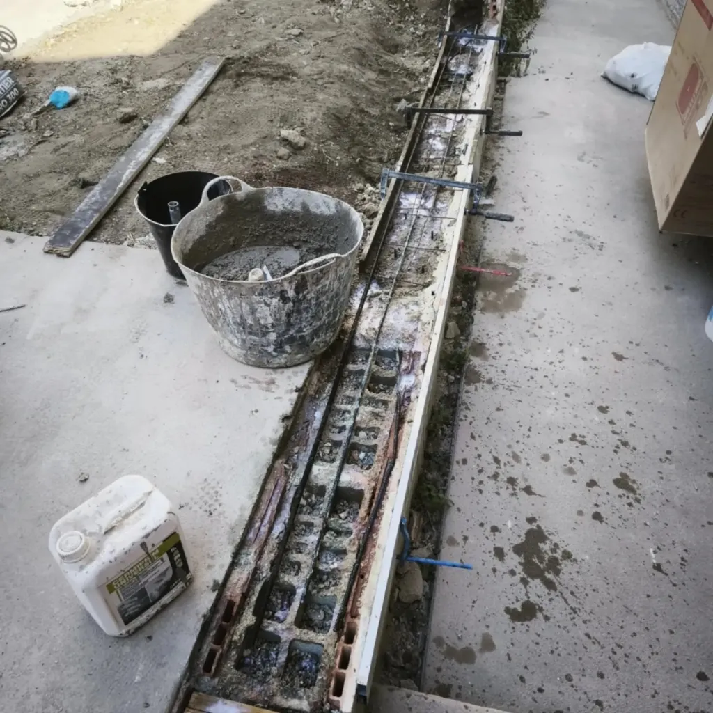 Chantier extérieur avec dalle béton, caniveau en cours de coulage, seau de mortier et bidon de produit.